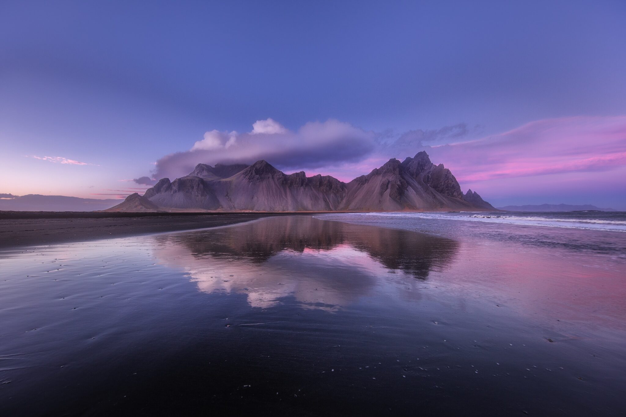 A purple and pink sky surrounds a mountain range in Iceland. The scene is reflected in the water that the mountains stand beside.