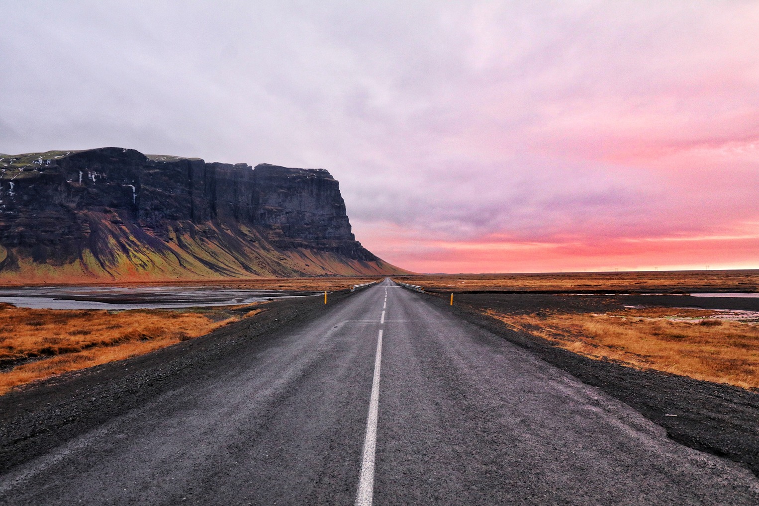 A long stretch of dark grey road surrounded by an orange landscape and cliffs with a pink cloudy sky during Womens Travel Network's Iceland tour for women